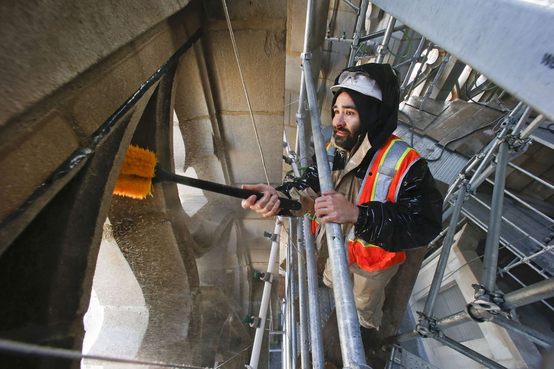 A construction worker in a high-visibility vest uses a brush to clean stone masonry while standing on scaffolding.