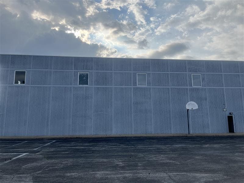 A gray, flat-walled building with four small square windows and a basketball hoop mounted on the right under a cloudy sky.