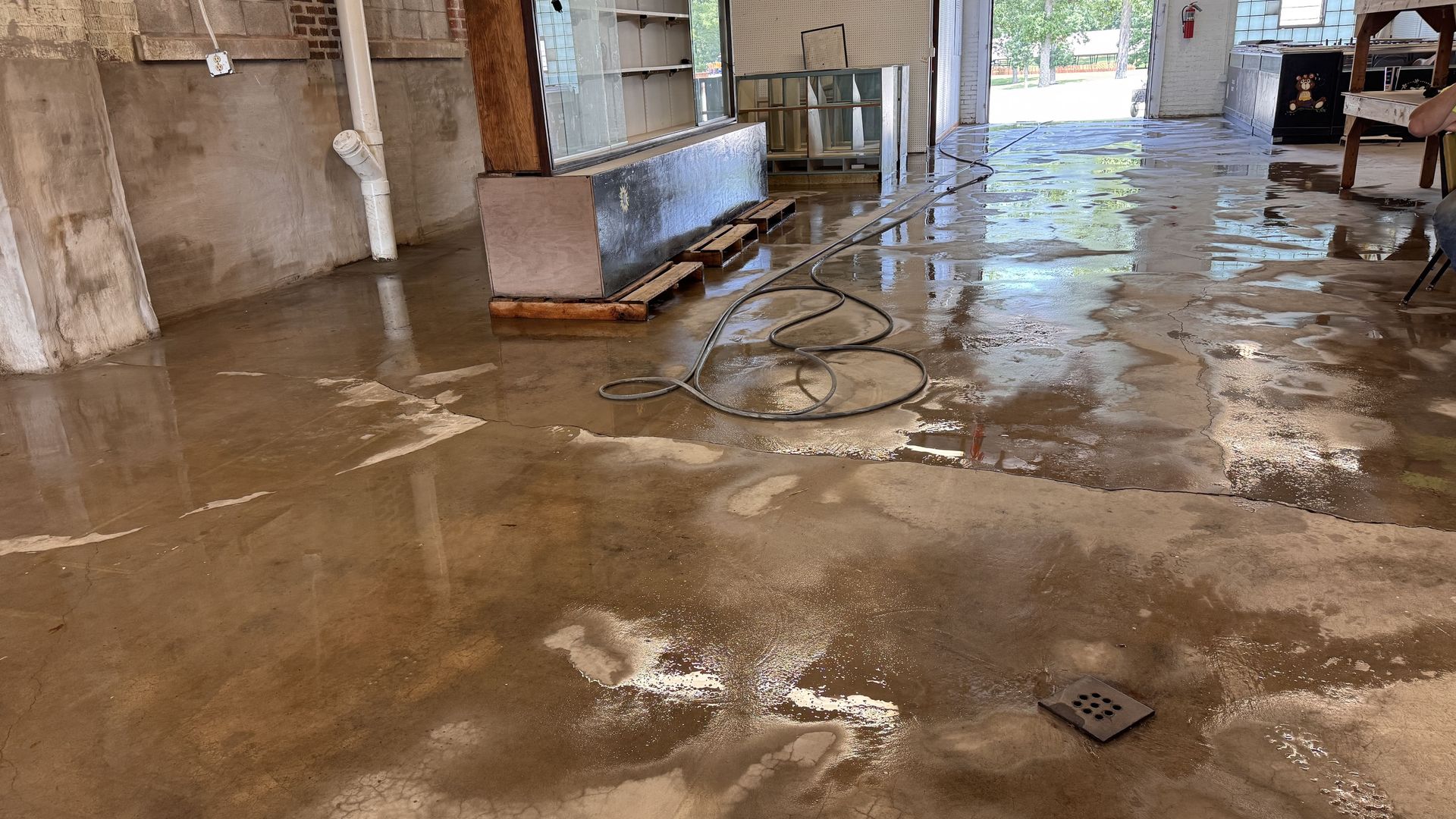 A wide-angle view of a damp, unfinished concrete floor in an indoor space with exposed walls and a coiled black hose.
