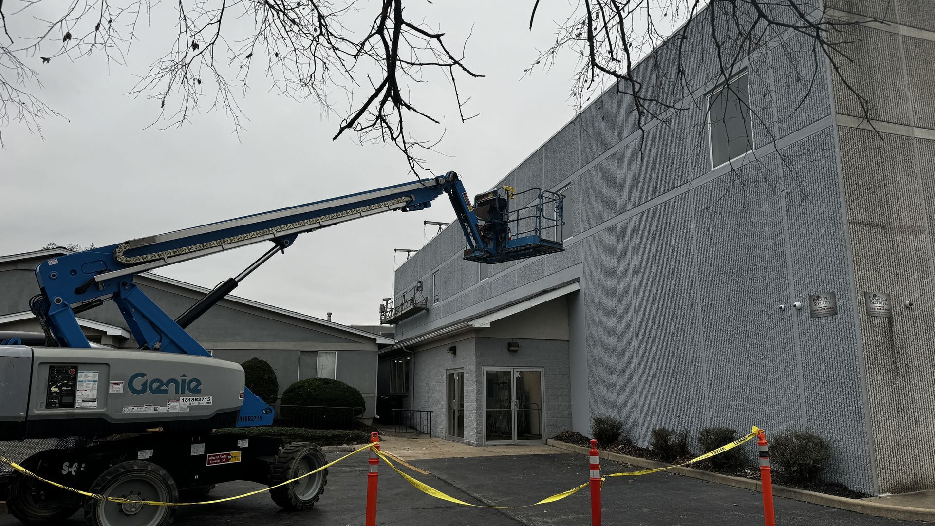 A blue Genie boom lift parked in a parking lot, extending its arm toward the side of a tall, gray building.
