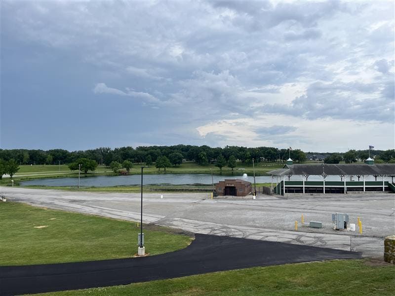 An open-air parking lot next to a pond and a covered outdoor structure under a cloudy sky.