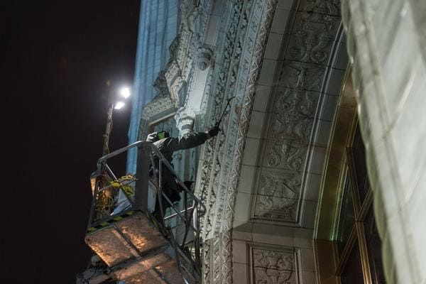 A person in a boom lift cleans the ornate stone archway of a building exterior at night under bright lighting.
