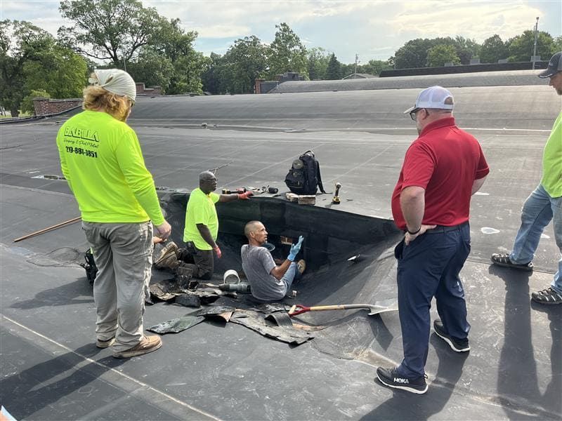 A construction team inspects a hole in a black flat roof, with one person working inside the opening.
