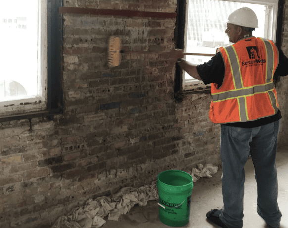 A worker in a high-visibility vest and hard hat paints an exposed brick wall near a window in an interior space.