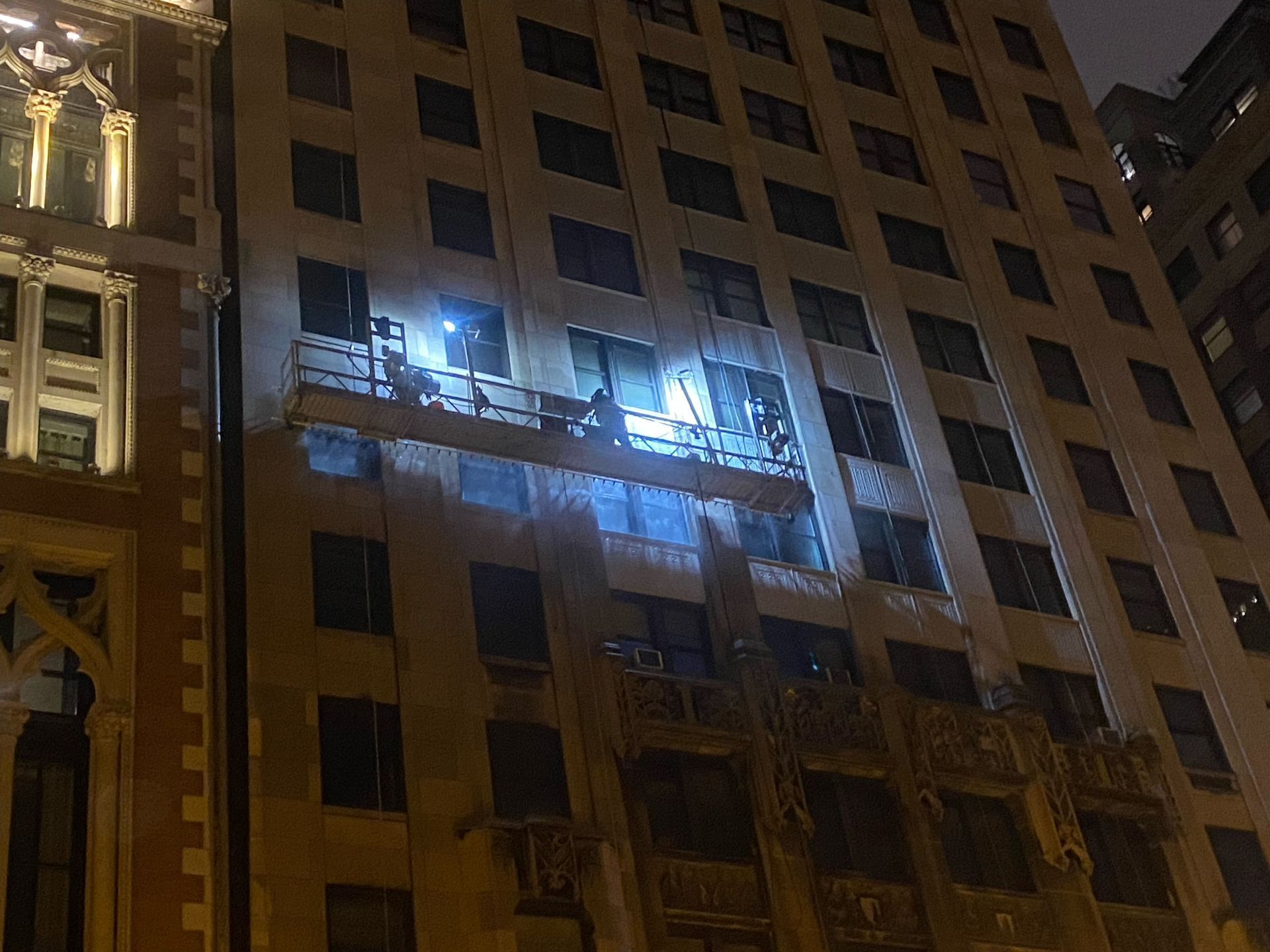 RestoreWorks crew performing water misting on Willoughby Tower at night, using suspended scaffolding with bright work lights. Specialists inside the building monitor for leaks to ensure no water infiltration. The nighttime work schedule helps minimize disruption to pedestrians below.
