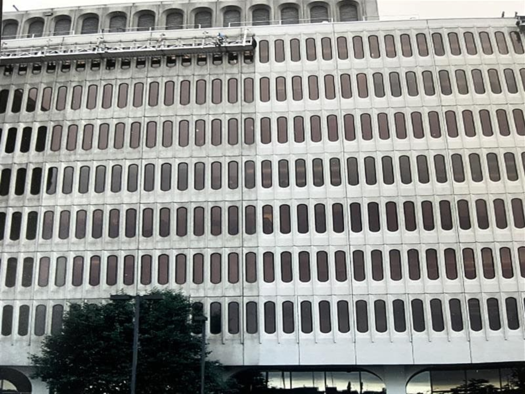 A large, Brutalist-style building with a repeating pattern of arched windows, half of which has been cleaned.