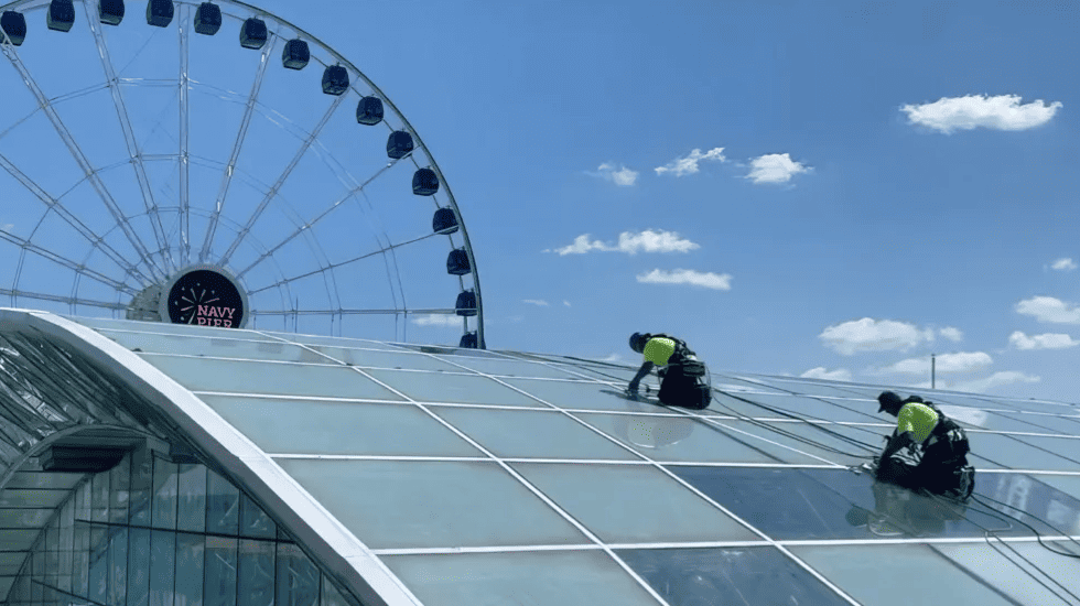 Two workers rope-access cleaning a curved glass roof with a ferris wheel in the background.
