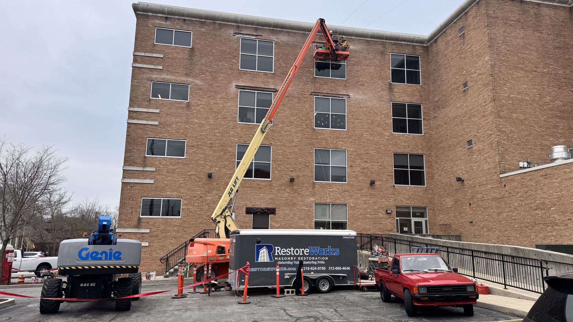 Construction workers on a cherry picker repairing windows on a brick building with dumpsters and a red car below