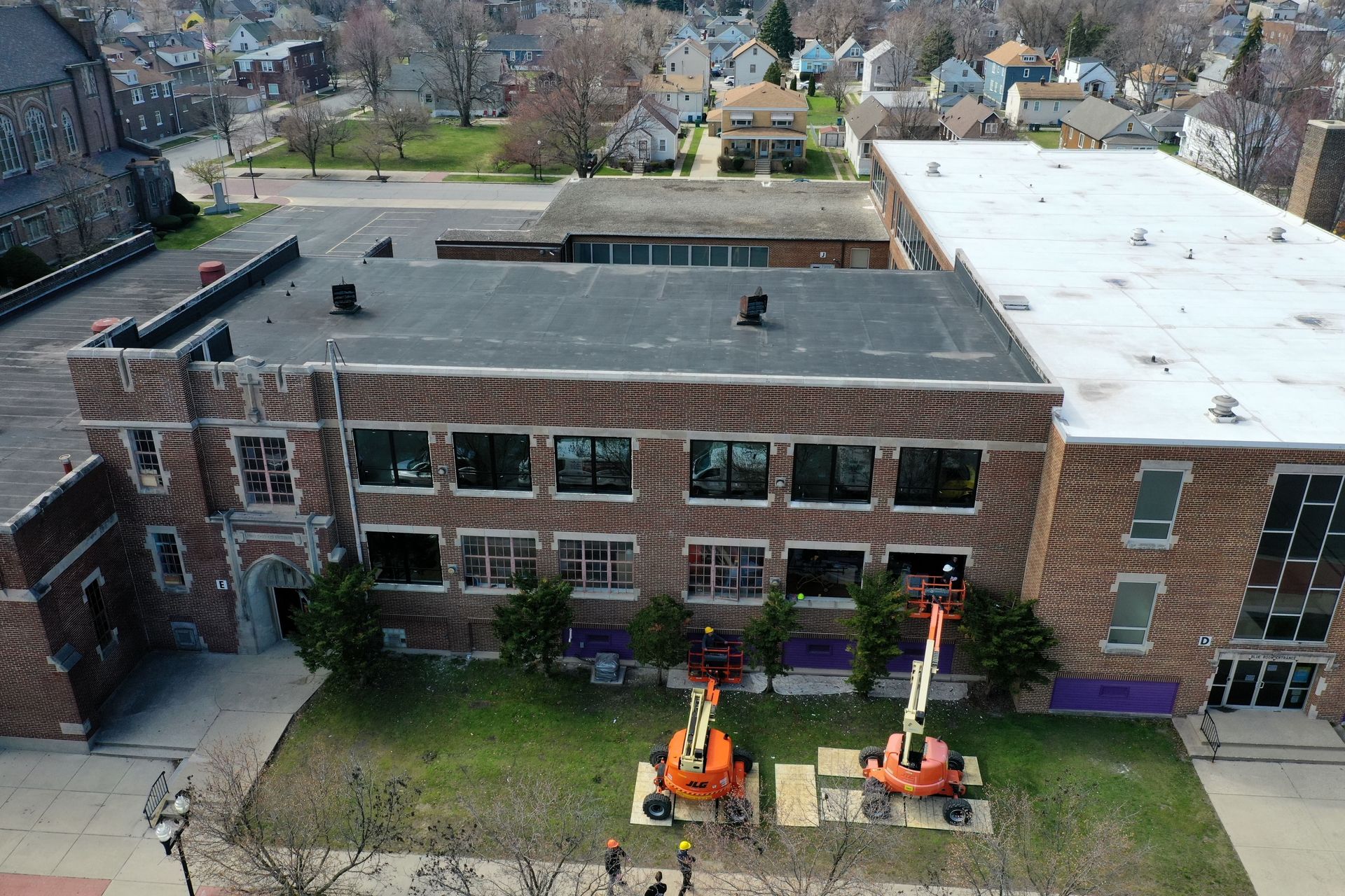 Aerial view of a brick school building with two orange cherry pickers parked in front on a grassy lawn.