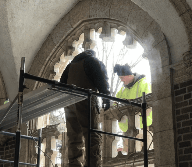 Two workers in safety gear on scaffolding inside a stone archway, inspecting or repairing a window