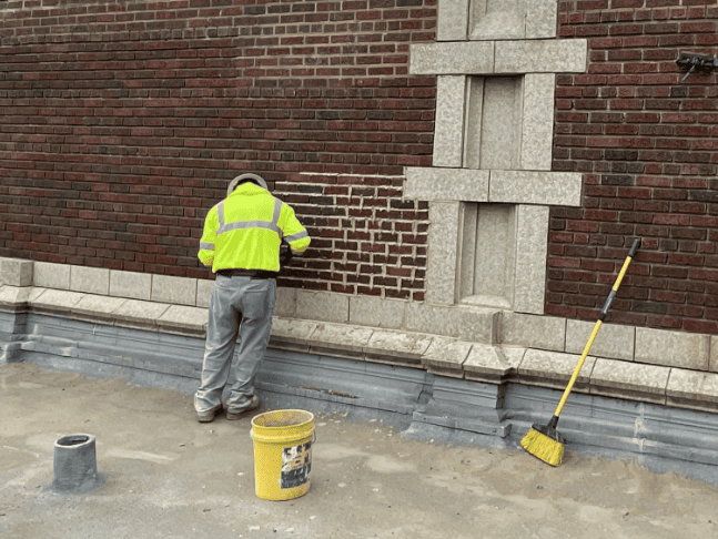 Worker in a neon jacket cleaning a brick wall beside a sidewalk, with tools and buckets nearby.