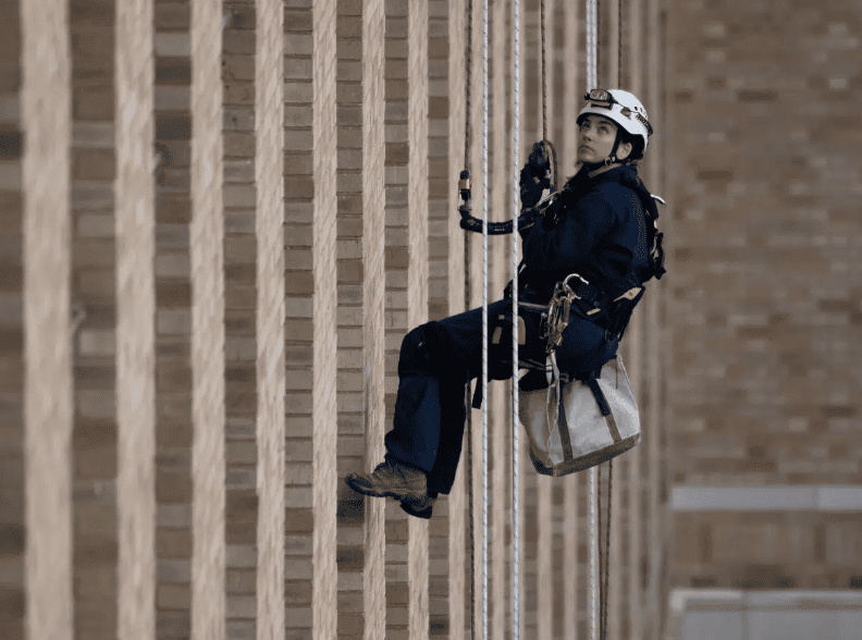 Worker suspended by ropes on a brick building facade, wearing a helmet and harness.