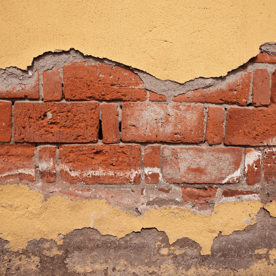 A deteriorating brick wall with severe spalling and peeling stucco. The outer layer of the bricks has crumbled away, exposing rough and eroded surfaces, while the yellow stucco coating is peeling off in large sections. White salt deposits and cracks in the mortar indicate moisture infiltration. This type of damage is commonly caused by water penetration and freeze-thaw cycles, leading to structural weakening over time.