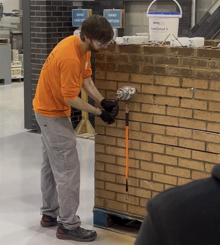A person in an orange shirt wears safety glasses and gloves while using a tool on a brick wall in a workshop.