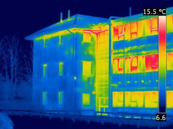  A thermal imaging scan of a multi-story building, displaying areas of energy loss through the building envelope. The image shows a color gradient, with blue representing cooler areas and red/yellow indicating heat loss. Warmer colors around windows, roof edges, and certain walls suggest poor insulation or air leakage. The scan highlights areas where energy efficiency improvements, such as better insulation or sealing, may be needed.