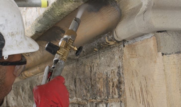 A worker in a hard hat and protective gloves uses a nozzle to spray a cleaning or restoration solution onto stone masonry.