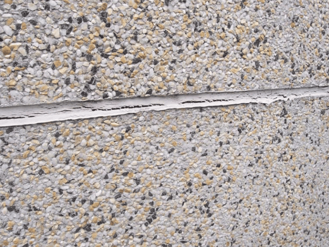 A horizontal gap in a textured, multi-colored aggregate concrete wall, showing a thin, peeling sealant strip.