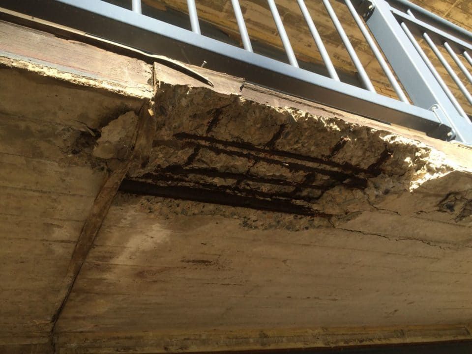 Damaged concrete balcony underside with exposed rebar and peeling, cracked edges
