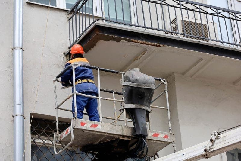 Worker in blue uniform and orange helmet on a lift, repairing a building’s exterior balcony area.