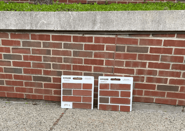 Two rectangular brick sample boards leaning against a red brick wall on a concrete sidewalk.