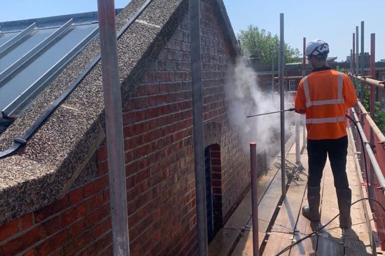 Worker in orange safety vest power-washing a brick wall on a rooftop construction site.