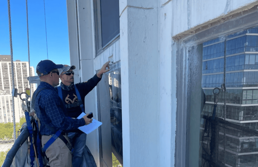 Two inspectors examine a white building exterior beside a window, one pointing at the wall.