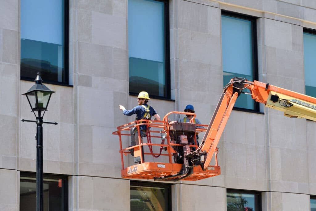 Two workers in a cherry picker beside a building, with one using an orange boom lift.