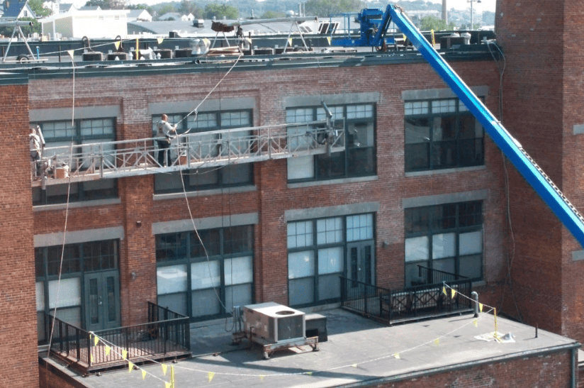 Rooftop view of a brick building with windows, railings, HVAC units, and a blue crane arm nearby