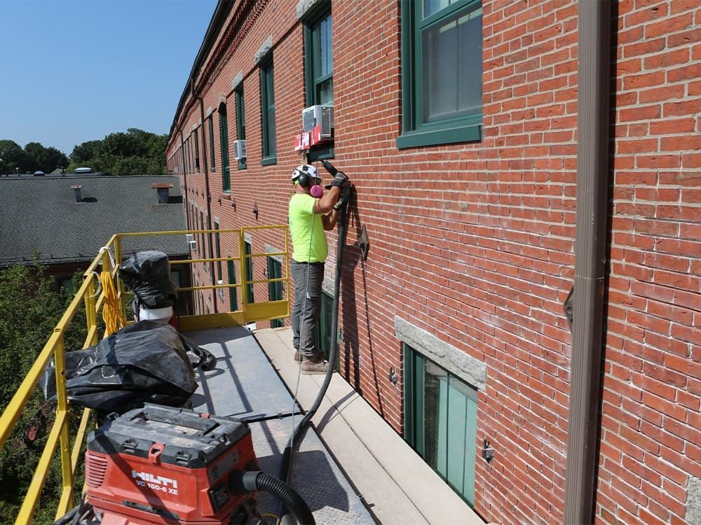Worker on a scaffold inspecting a brick building wall beneath green-trimmed windows.