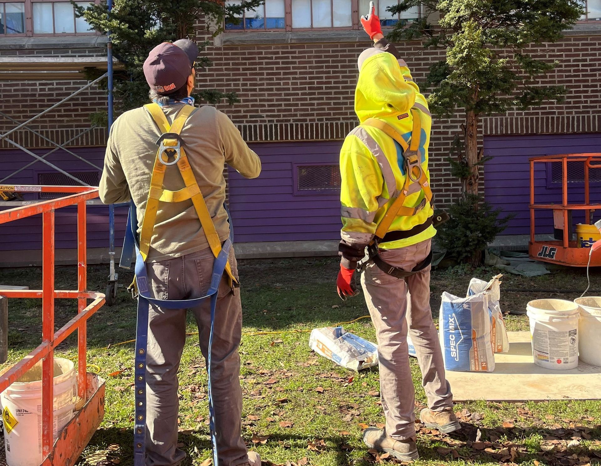 Two construction workers in safety harnesses stand outdoors by a brick building, one pointing toward the wall.