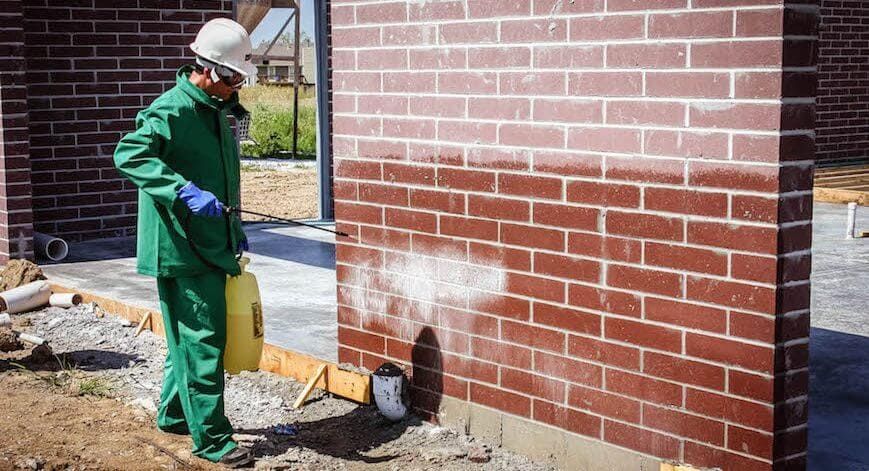 A person in protective clothing and a hard hat sprays a chemical treatment onto a brick wall at a construction site.