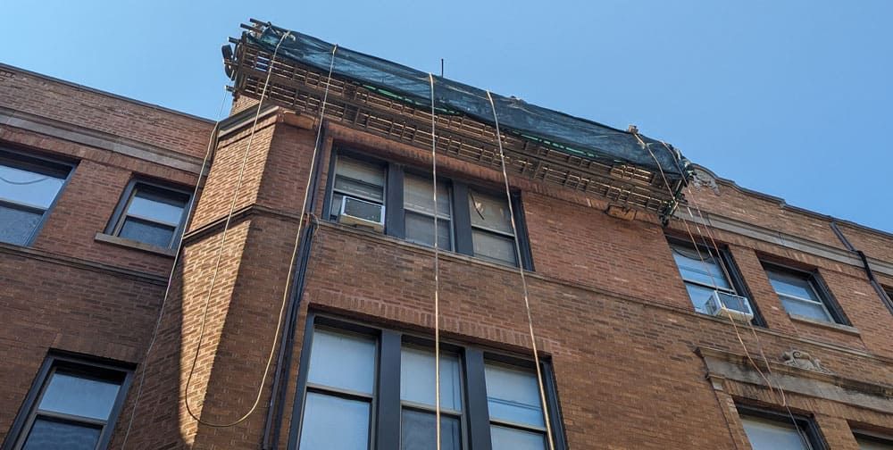 A low-angle view of a brick apartment building facade with a safety net draped across the top edge under a blue sky.