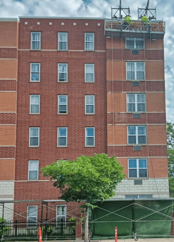 Red brick apartment building behind a green tree and fenced sidewalk under cloudy sky
