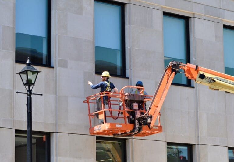 Two workers in an orange cherry picker repairing a building facade beside a streetlamp.