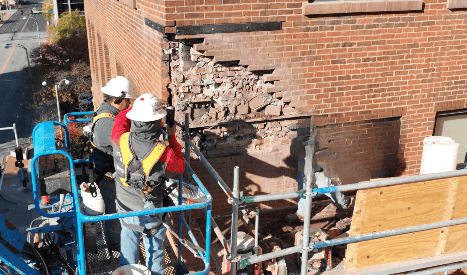 Workers on blue lifts repair a damaged brick wall beside a building.