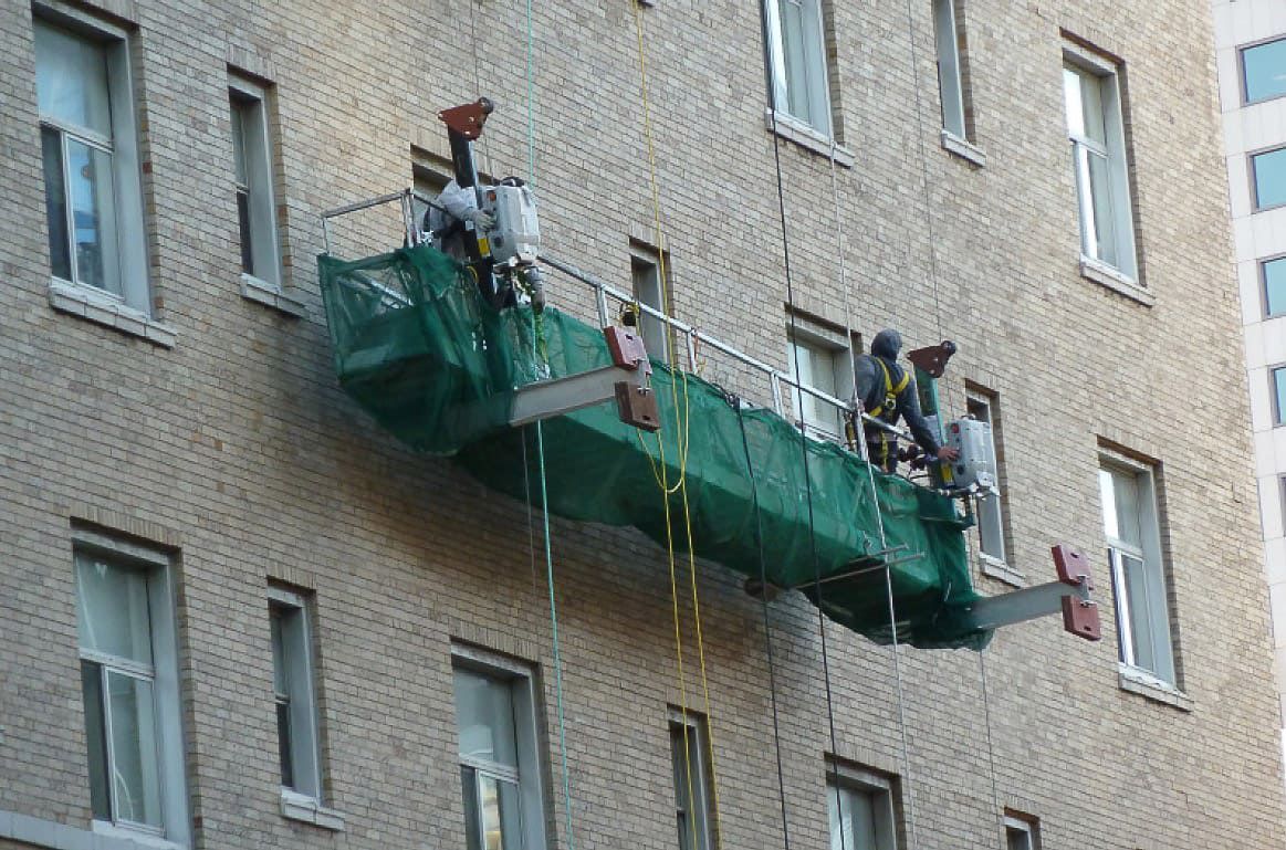 Workers on a green suspended platform painting the exterior wall of a brick apartment building.
