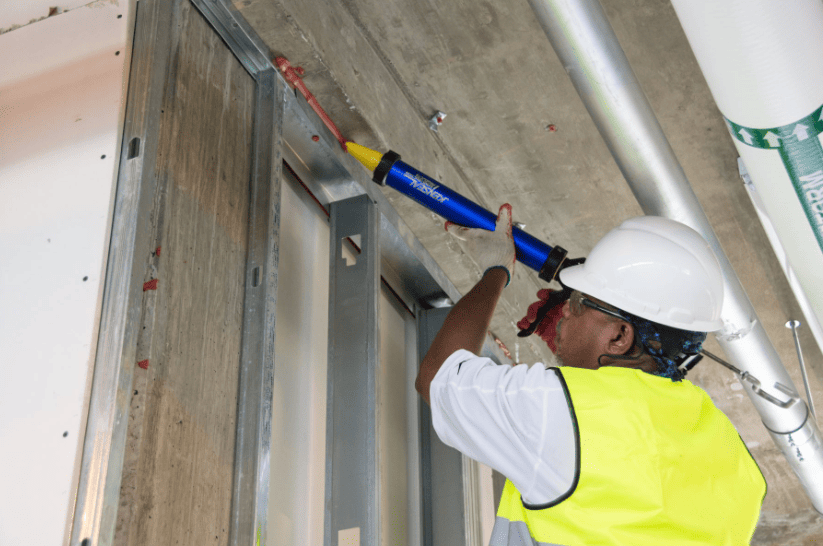 Worker in hard hat and safety vest installing an overhead fixture indoors with a power tool