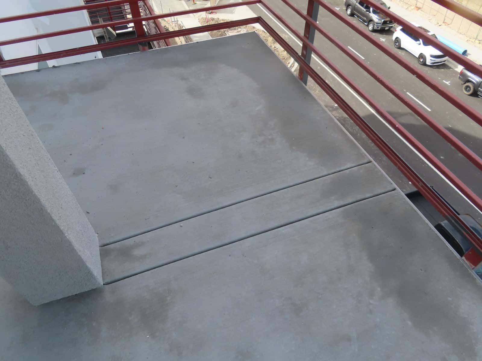 Concrete balcony with red railing and a closed floor hatch overlooking a street below