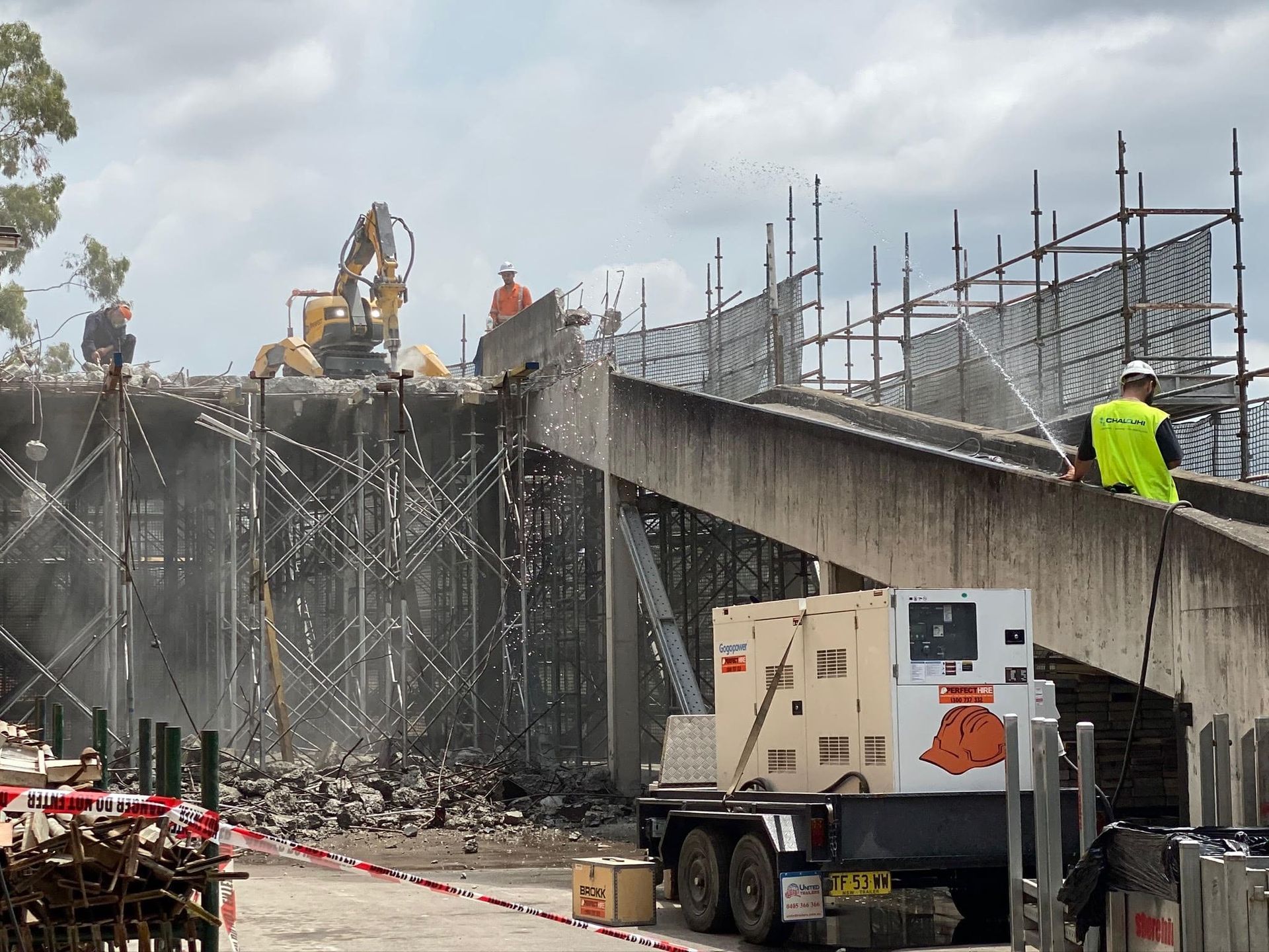 Construction workers on scaffolding beside a partially demolished concrete structure and excavator.
