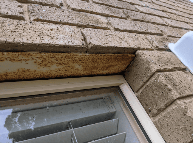 Close-up of a tan stone exterior wall above a window with rust-stained metal trim and white blinds.
