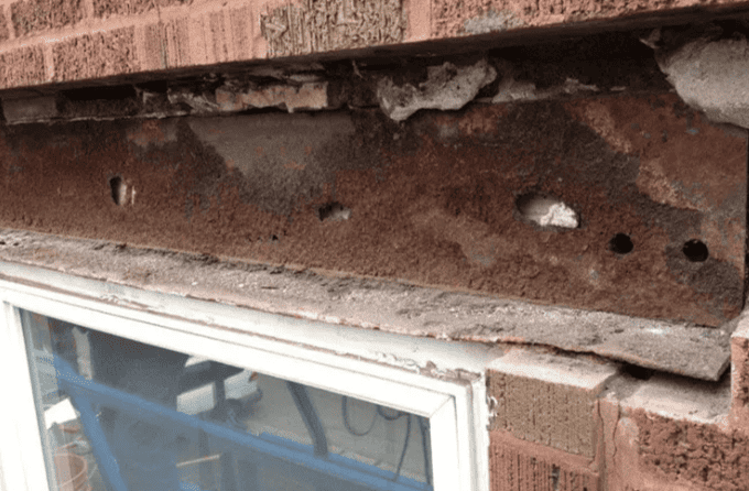 Rusty steel lintel above a white-framed window in a brick wall with visible corrosion and holes