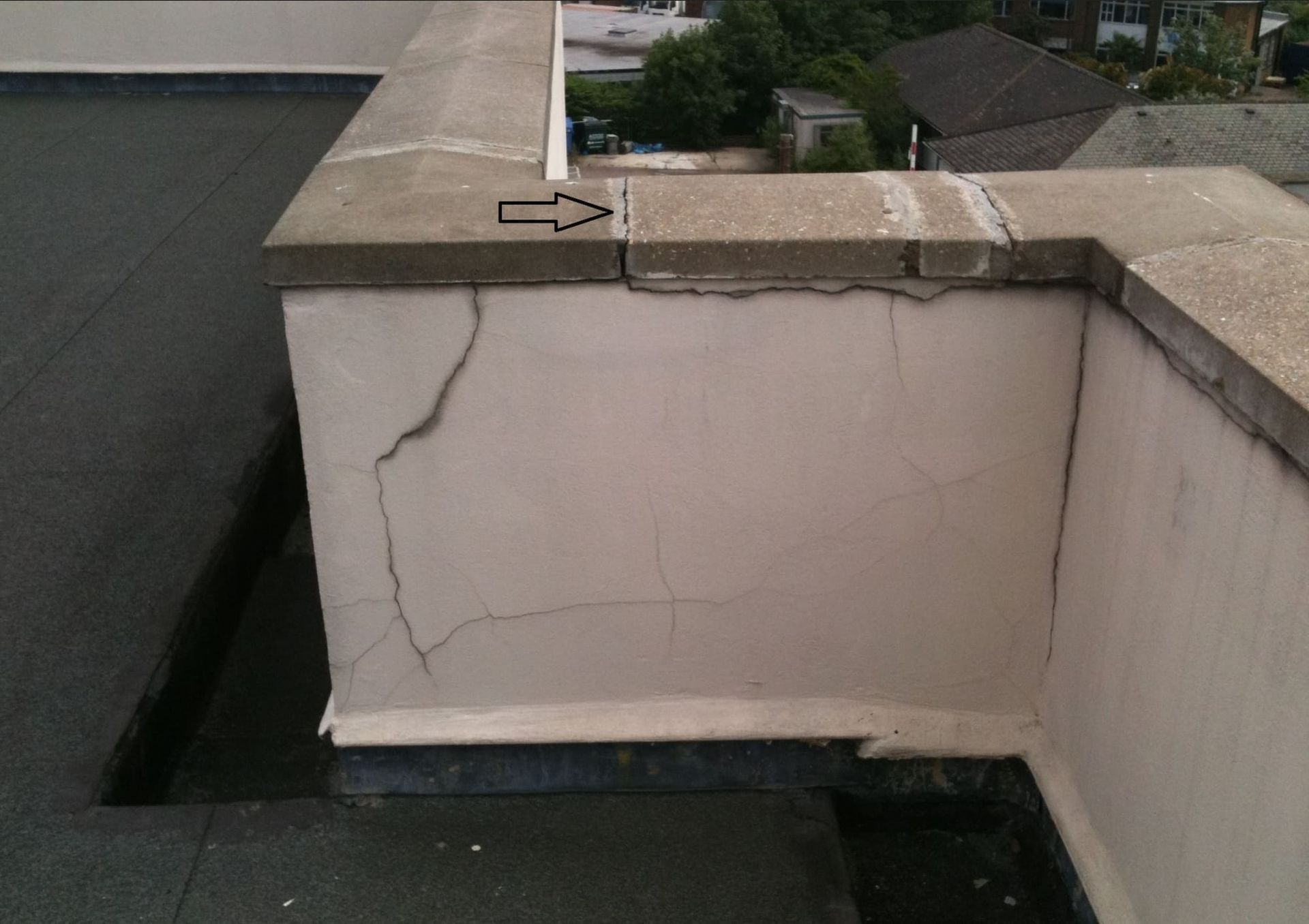 A roof parapet wall shows cracks in the stucco finish and a gap between the stone capstones, marked by a black arrow.