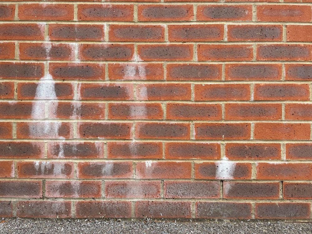 Red brick wall with white spray-painted graffiti and a small exposed pipe near the bottom right.