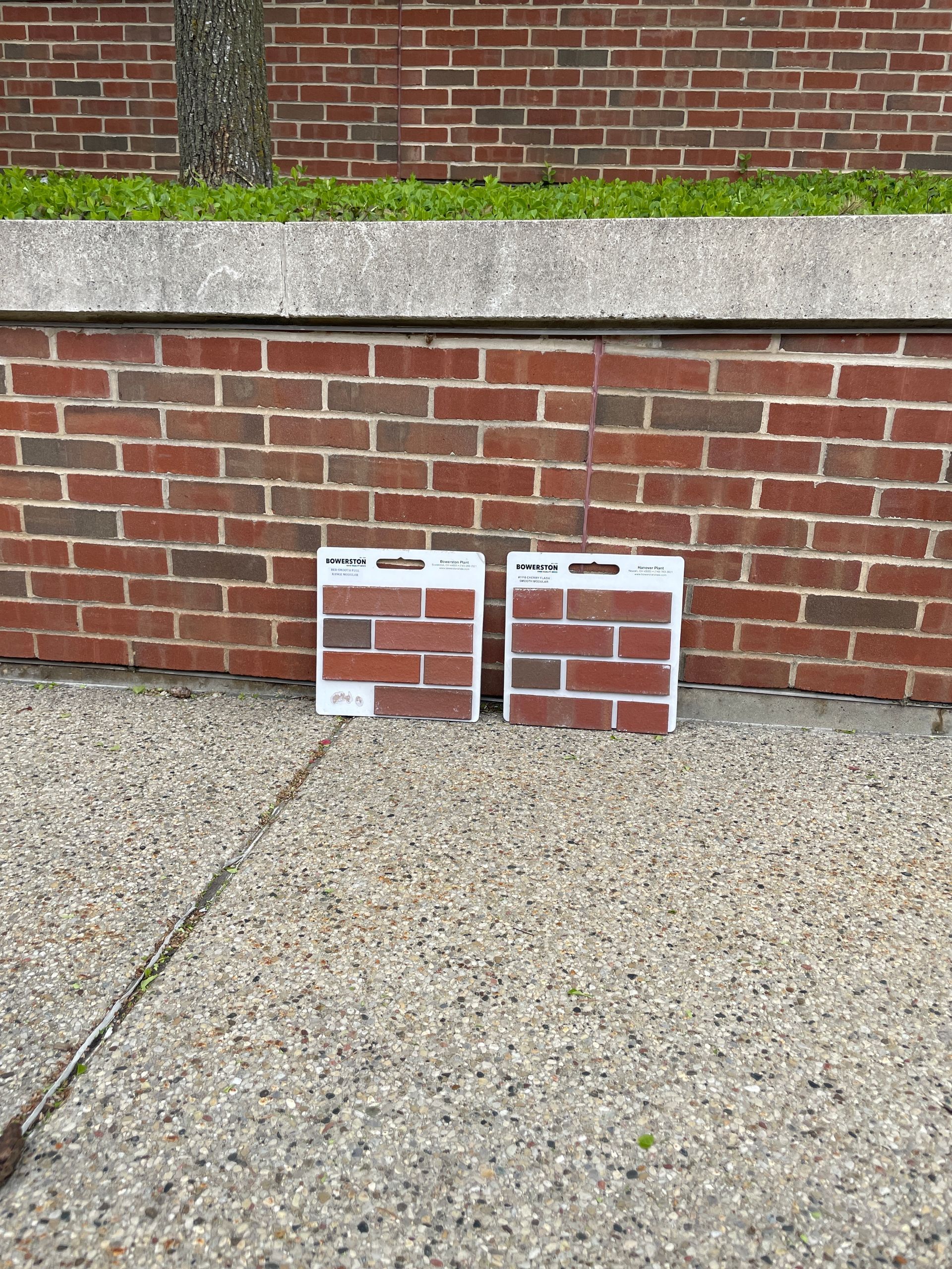 Two brick samples are sitting on the sidewalk in front of a brick wall.