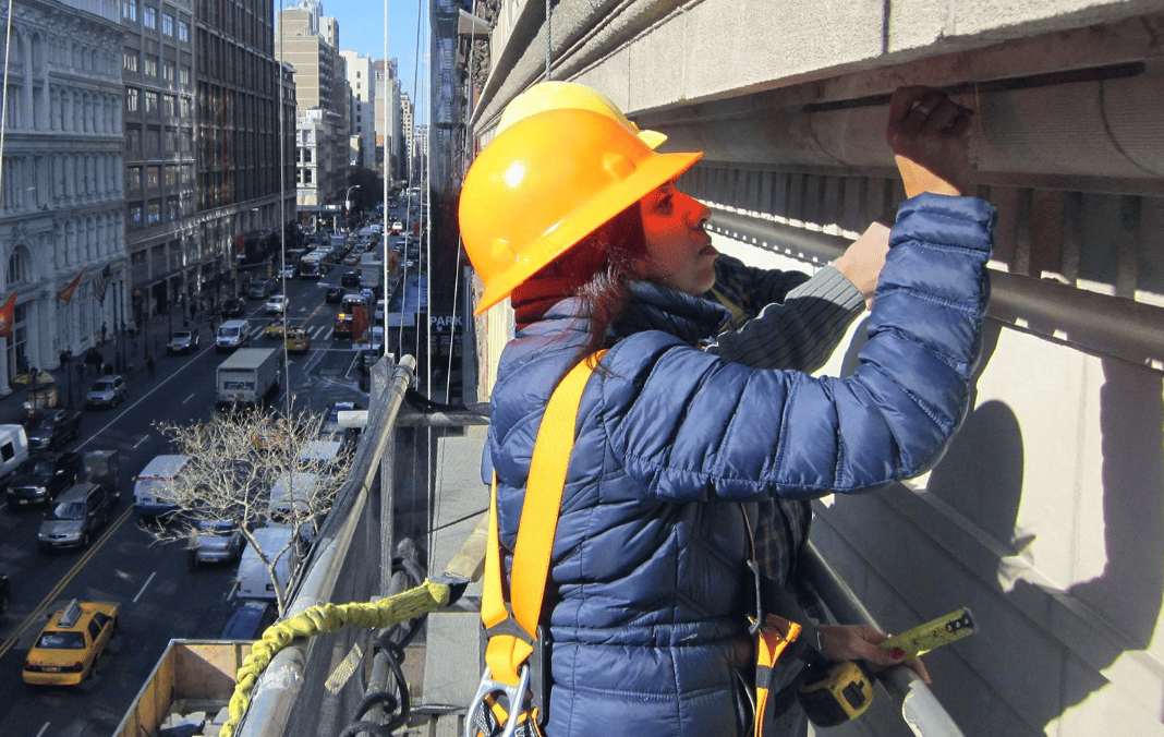 Worker in orange hard hat and blue jacket repairing building facade on a suspended platform above a city street