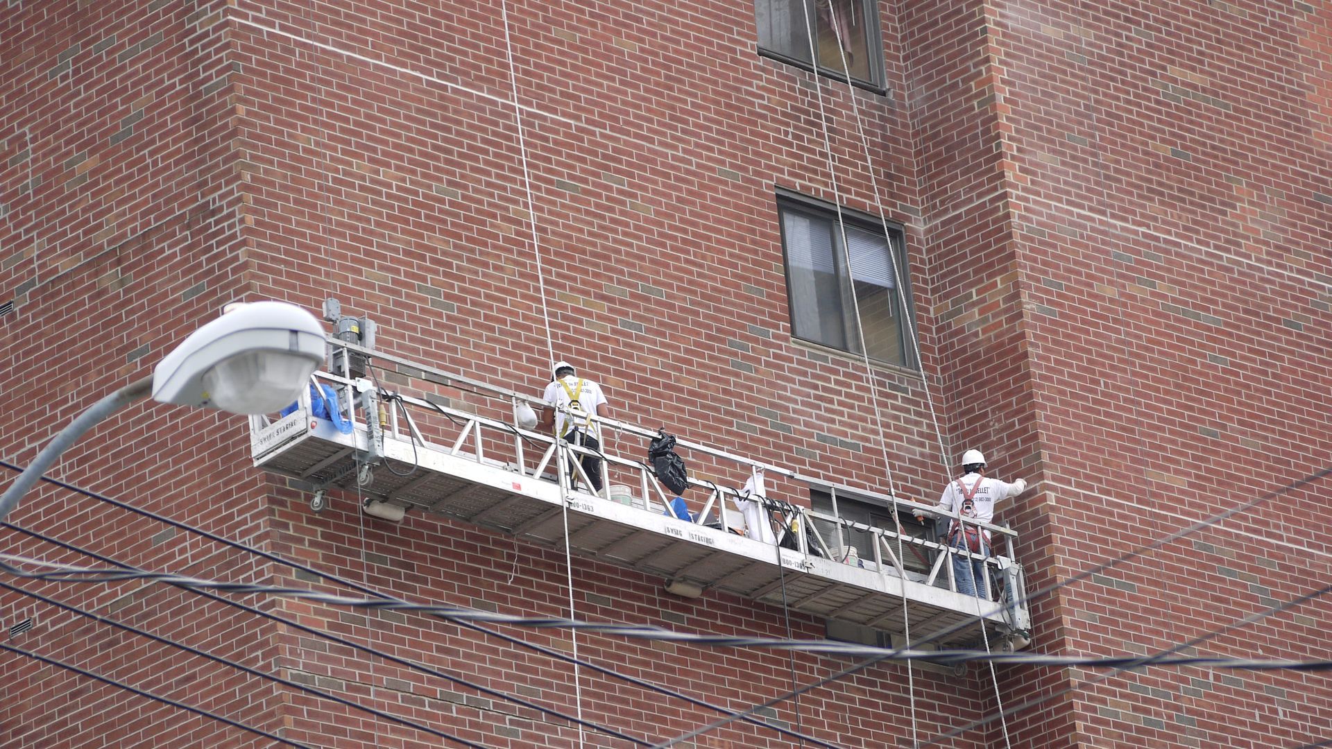 Workers on a suspended platform outside a brick building, installing or repairing the facade.