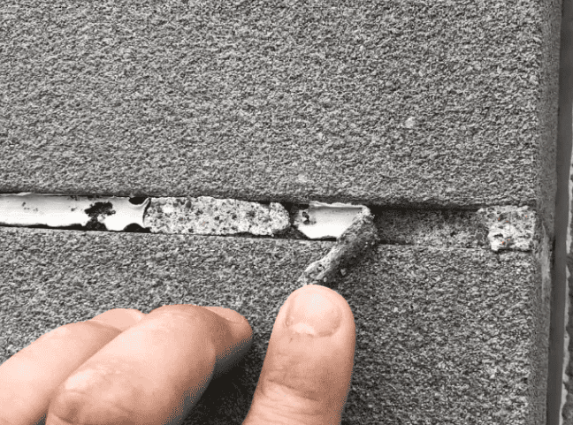 Finger pointing at a cracked gray wall with peeling white paint in a damaged seam