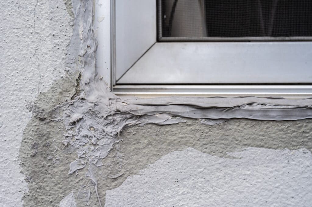 Damaged stucco wall around a white window frame with peeling plaster and cracks