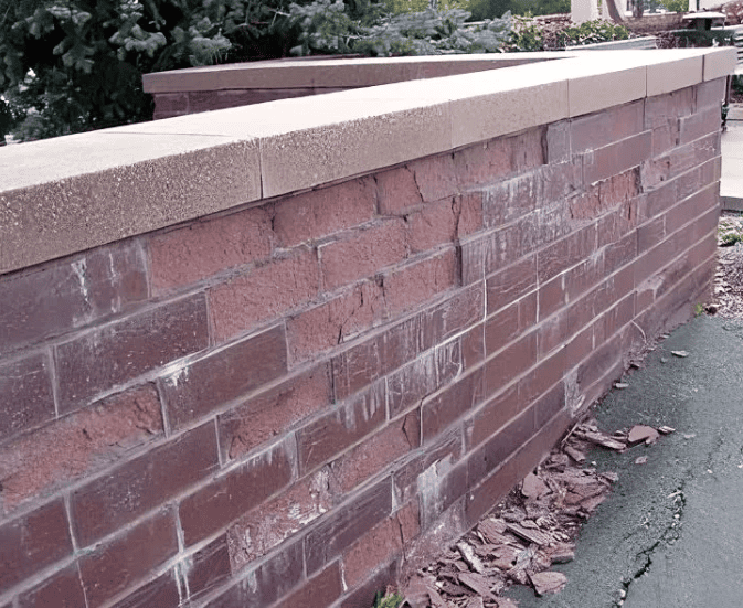Brick retaining wall beside a sidewalk, topped with tan stone and scattered dry leaves.