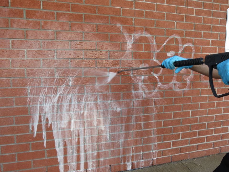 Technician using a pressure washer and cleaning solution to remove graffiti from a red brick commercial building during professional masonry cleaning.